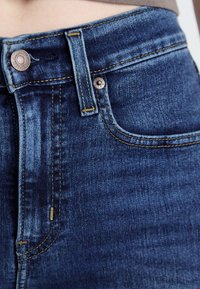 Temno modri jeans z klasičnim krojem, opremljeni z kovinsko zaponko, rumenim šivom in gladko, rahlo zbledelo teksturo.