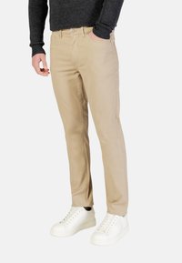Pantalones de algodón beige con un ajuste entallado, diseño de cinco bolsillos y cierre de botón, combinados con zapatillas blancas. Tejido texturizado con un acabado suave.