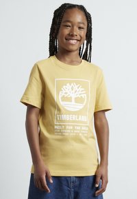Een korte mouwen gele katoenen t-shirt met een wit Timberland-logo en tekst. Gepaard met blauwe denim korte broeken. Effen achtergrond.