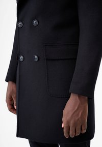 Dubbelt knäppt svart ullkappa med stora svarta knappar, en sidficka och en mjuk textur som framhäver dess strukturerade design.