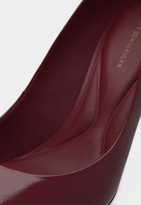 Κοντινό πλάνο ενός μπορντό δερμάτινου γυναικείου τακουνιού που δείχνει την επενδυμένη εσωτερική σόλα με το λογότυπο Tommy Hilfiger.