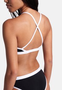 Svart bikinitopp med vita accenter, med en design som korsar ryggen och justerbara remmar. Slät, elastisk tyg med minimal täckning.