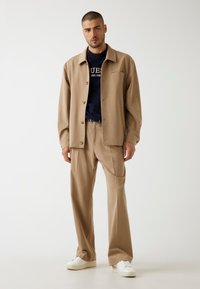 Beige lichtgewicht button-up jack over een navy shirt, gecombineerd met op maat gemaakte beige broeken en witte sneakers. Minimalistisch ontwerp, subtiele texturen.