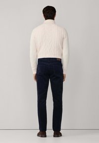 Krémový sveter s pleteným vzorom a vysokým golierom, námornícke nohavice a hnedé topánky. Sveter obsahuje textúrované vzory; nohavice sú slim-fit.