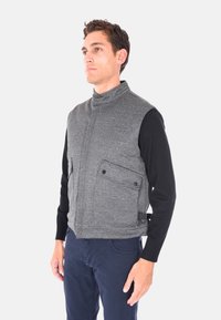 Gilet grigio senza maniche con collo alto, tessuto testurizzato, due tasche frontali con chiusure a scatto e cintura laterale regolabile; indossato sopra una maglietta nera a maniche lunghe.