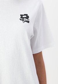 T-shirt en coton blanc à manches courtes avec un petit motif graphique noir sur la partie gauche de la poitrine. Texture douce.