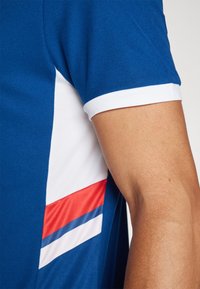 Polo bleu avec col et manches blanches, présentant des détails de panneaux latéraux en rouge et blanc, mettant en avant un tissu texturé et un design sportif.