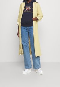 Żółty trencz na granatowej bluzie z kapturem z logo, zestawiony z rozkloszowanymi niebieskimi dżinsami i białymi trampkami. Gładka tkanina, casualowy design.