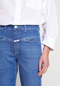 Personne portant un jean en denim bleu et une chemise blanche à boutons, la main droite détendue le long du corps.