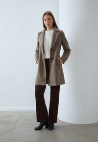 Manteau en laine beige avec un col cranté, deux poches avant et deux boutons, associé à un pantalon en velours côtelé marron foncé et des bottes noires à talons.