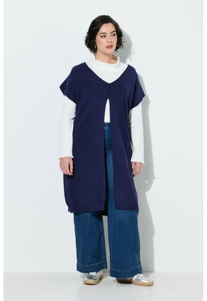 Kvinde med kort mørkt hår iført en lang marineblå vest over en hvid top og brede blå jeans, stående mod en ensfarvet lys baggrund.