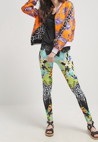 Giacca bomber arancione con motivi floreali, indossata sopra un top nero. Leggings colorati con design geometrici e tropicali. Sandali neri.
