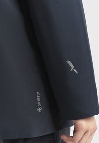 Námořnická modrá Gore-Tex bunda zblízka; má hladkou strukturu s logem a emblémem na rukávu, což ukazuje voděodolný materiál a design.