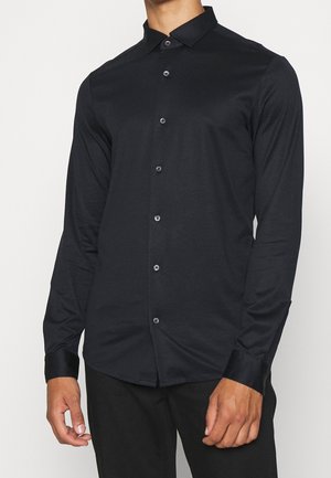 Man wearing a fitted long-sleeve black button-up shirt with collar, standing against a plain light background.