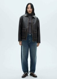 Veste en simili cuir marron avec des coutures prononcées, deux poches avant, col roulé gris et jean bleu ample associé à des chaussures foncées.