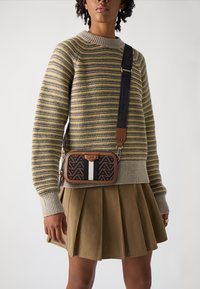 Brun mönstrad axelväska med svartvit rand, fäst vid ett svart band. Modellen bär en randig tröja och en plisserad kjol.