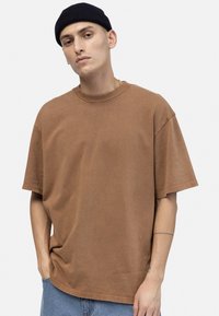 Young man wearing a brown oversized t-shirt, blue jeans, and a black beanie, standing with one hand in pocket against a plain background.