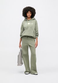 Γυναίκα με σγουρά μαλλιά, φορώντας ελιά χρώματος φούτερ με κουκούλα Nike Shox και φαρδιά παντελόνια, κρατώντας γκρι τσάντα ώμου σε λευκό φόντο.