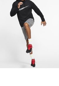 Zwarte hoodie met een wit logo, grijze shorts, witte en neon gele sokken, en zwarte sneakers met rode veters, in een actiepose midden in een stap.