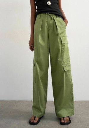 Person iført løse olivengrønne cargobukser med store sidelommer, en sort ærmeløs top, sorte sandaler og en sølv vedhæng halskæde.