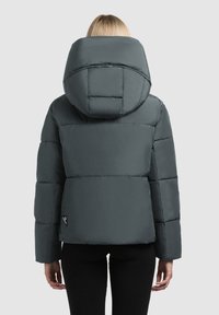 Kvinde iført en mørkegrå polstret jakke med en stor hætte, set fra ryggen mod en lys ensfarvet baggrund.