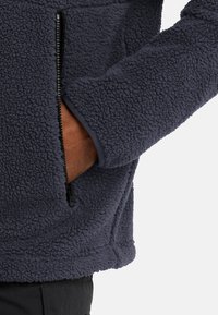 Giacca in pile blu navy con finitura tessurizzata, caratterizzata da una tasca con zip e un tessuto morbido e confortevole. I polsini hanno un bordo di colore uniforme.