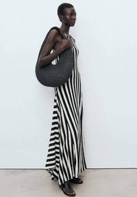 Striped maxi dress in black and white fabric; features vertical stripes. Model carries a textured, dark woven bag and wears flat sandals.