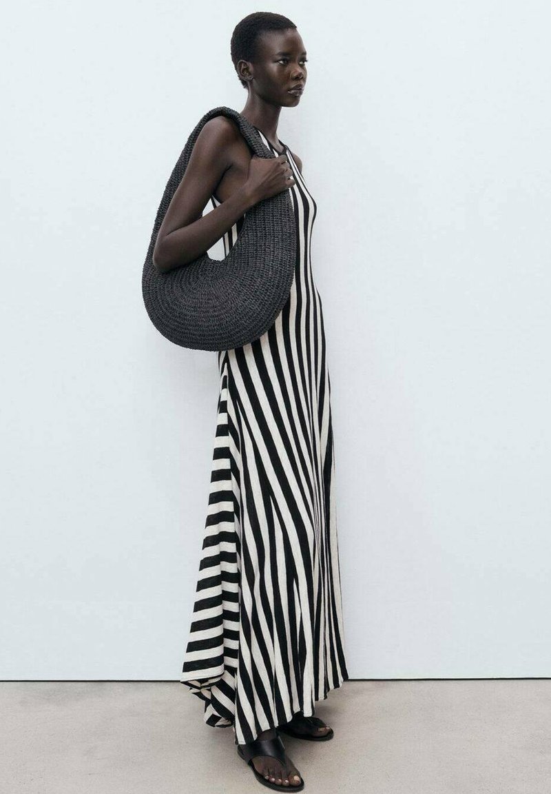Striped maxi dress in black and white fabric; features vertical stripes. Model carries a textured, dark woven bag and wears flat sandals.