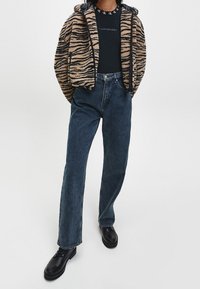 Brązowa i czarna, paskowana kurtka z sztucznego owczego futra z zamek błyskawicznym, w zestawieniu z ciemnoniebieskimi, wysoko taliowanymi dżinsami i czarnymi butami.