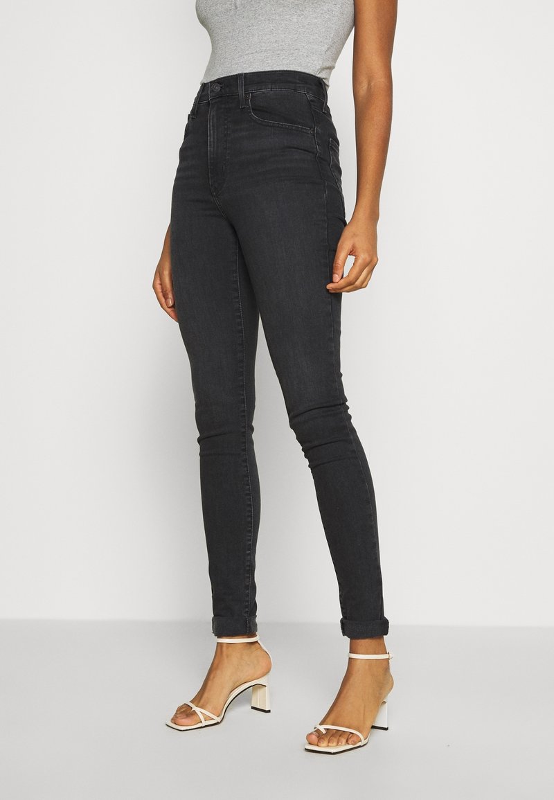 Jean skinny noir taille haute avec une coupe slim, doté de passants de ceinture, poches avant et ourlets retroussés, associé à des talons blancs à lanières.