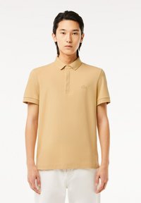 Beige pikétröja med mjuk textur, knappad krage och korta ärmar. Har en liten broderad logotyp på bröstet.