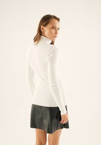 Suéter blanco ajustado de punto acanalado con cuello alto y mangas largas, combinado con una mini falda de cuero negra, destacando el contraste de texturas y diseño.