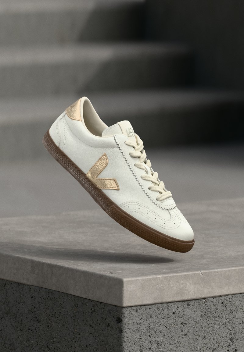 Baskets en cuir blanc avec accents dorés, semelle en caoutchouc texturée et laçage classique. Le côté présente un logo distinctif en forme de "V" dans une teinte beige.