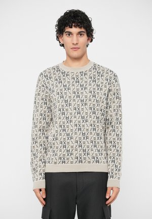 Jeune homme aux cheveux bouclés portant un pull beige à motifs et un pantalon cargo noir, se tenant devant un fond blanc uni.