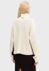 Donna bionda che indossa un maglione oversize in crema a coste con collo alto e maniche lunghe, abbinato a una gonna nera a coste con spacco sul retro, di spalle.