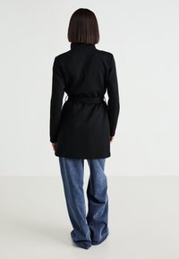 Zwarte coat met een hoge kraag en een getailleerde pasvorm, gecombineerd met losse blauwe denim jeans. De stof lijkt glad met minimale textuur.