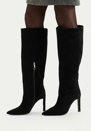 Schwarze kniehohe Stiefel aus Wildleder mit spitzen Zehen und Stöckelabsätzen, getragen mit schwarzen Strumpfhosen und einem grauen Rock.