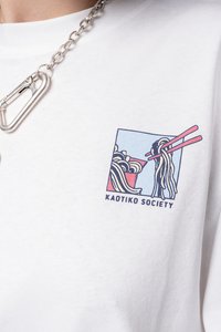 Valkoinen puuvillapaita, jossa on brodeerattu malli nuudeleista ja syömäpuikoista. Suunnitelmassa on rohkeita värejä ja teksti "KAOTIKO SOCIETY."