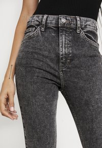 Primer plano de una persona que lleva puestos unos pantalones vaqueros de talle alto, negros y desgastados, y una camiseta negra de canalé, con un tatuaje de flecha en el antebrazo izquierdo.