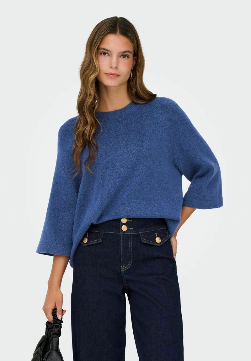 Femme aux longs cheveux ondulés portant un pull bleu ample, un jean taille haute foncé avec des boutons dorés, tenant un sac à main noir froncé.