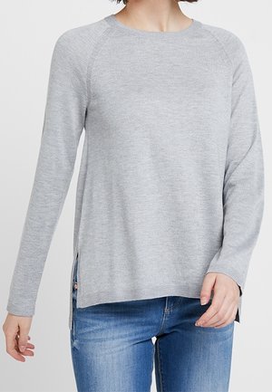 Žena oblečená v světle šedém svetru s dlouhými rukávy a postranními rozparky, v kombinaci s modrými džínami, stojí před jednobarevným pozadím.