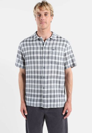 Homme blond portant une chemise à manches courtes à carreaux gris et blanc boutonnée et un pantalon foncé, debout devant un fond clair uni.