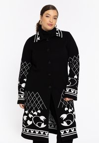 Model trägt einen schwarzen Mantel mit weißen floralen und geometrischen Mustern, der einen hohen Kragen und einen Knopfverschluss hat, vor einem schlichten weißen Hintergrund.