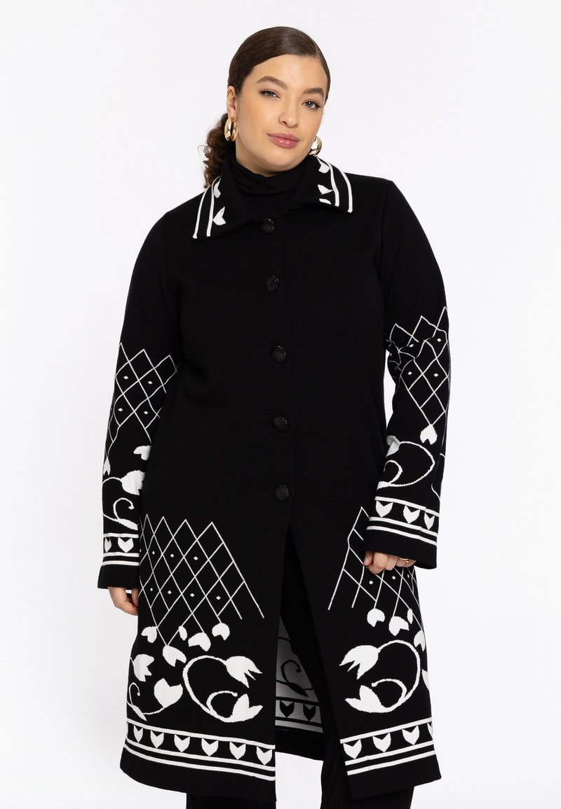 Model trägt einen schwarzen Mantel mit weißen floralen und geometrischen Mustern, der einen hohen Kragen und einen Knopfverschluss hat, vor einem schlichten weißen Hintergrund.