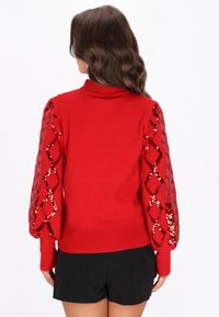 Czerwona sweter z prążkowanym dekoltem i bufiastymi rękawami wyszywanymi cekinami, zdobiony wzorem w diamenty. Materiał wydaje się miękki i teksturowany.