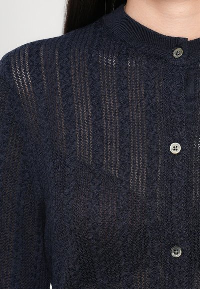 Laivastonsininen neulecardigan, joka on valmistettu tekstiilipinnasta, jossa on pystysuoria kuvioita. Siinä on pyöreä kaulus ja yksi nappi kiinnitykselle.