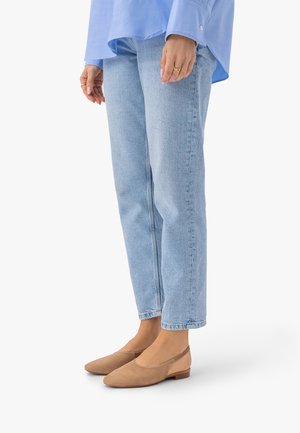 Osoba ubrana w jasnoniebieskie dżinsy i beżowe zamszowe baleriny, stojąca z rękami luźno opuszczonymi wzdłuż ciała, na jednolitym białym tle.