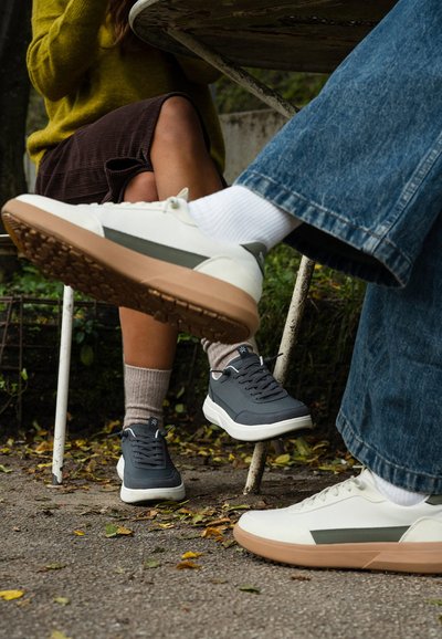 Sneakers bianchi con dettagli verdi e suole beige accanto a sneakers grigio scuro con suole bianche. Entrambi presentano texture lisce e materiali morbidi.