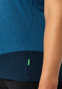 Chemise bleue à manches courtes avec une texture chinée, présentant un panneau inférieur contrastant en bleu foncé et une étiquette de marque verte sur l'ourlet.