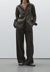 Chemise à boutons vert olive foncé associée à un pantalon ample assorti. Silhouette décontractée, tissu doux et poignets prolongés. Sandales noires.